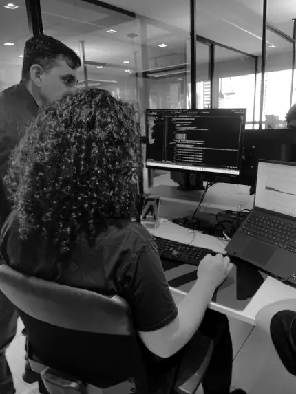 Equipe técnica colaborando enquanto escreve códigos, com computadores, notebooks e telas exibindo linhas de programação em um ambiente de trabalho.